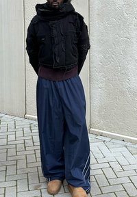 Musta lyhyt huppari, jossa on purkautuneet reunat, yhdistettynä löysiin tummansinisiin housuihin, joissa on valkoiset sivupinnat ja vaaleanruskeat kengät. Rakenne on tekstuuriltaan kerroksellinen.