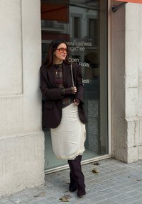 Femme en blazer marron, haut à carreaux, jupe crème et bottes hautes foncées, appuyée contre un mur à côté d'une porte vitrée affichant les horaires du magasin.