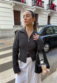 Giacca nera con zip e colletto, indossata sopra una gonna bianca. Collana di perle e orecchini a cerchio in tono argentato. Borsa nera con un portachiavi a forma di orsetto.