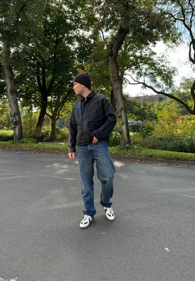 Chaqueta bomber negra, jeans azules y zapatillas blancas con detalles en negro. De pie al aire libre entre árboles con la luz del sol filtrándose a través de las hojas.