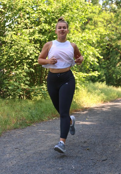 Corredor con una camiseta sin mangas blanca de malla y leggings negros. Zapatillas deportivas grises en un camino de grava rodeado de vegetación.
