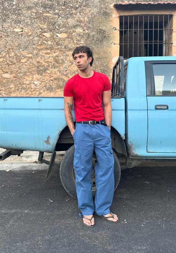 Giovane uomo con tatuaggio, indossa una camicia rossa, pantaloni blu e sandali, appoggiato a un pick-up blu accanto a un muro di pietra.