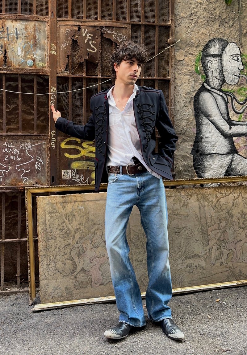 Black military-style jacket with decorative hardware, white shirt, faded blue jeans, and black shoes. Urban graffiti backdrop.