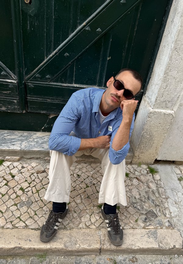 Mann mit Sonnenbrille, blau gestreiftem Hemd und cremefarbenen Hosen sitzt auf einem Kopfsteinpflaster-Gehweg und lehnt an einer dunkelgrünen Tür.