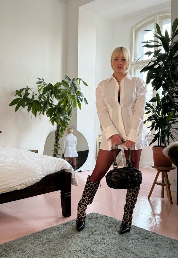 Biała, oversize koszula-dress, czarna torba na ramię z metalowymi dodatkami, kozaki w panterkę do kolan, wnętrze z roślinami i okrągłym lustrem.