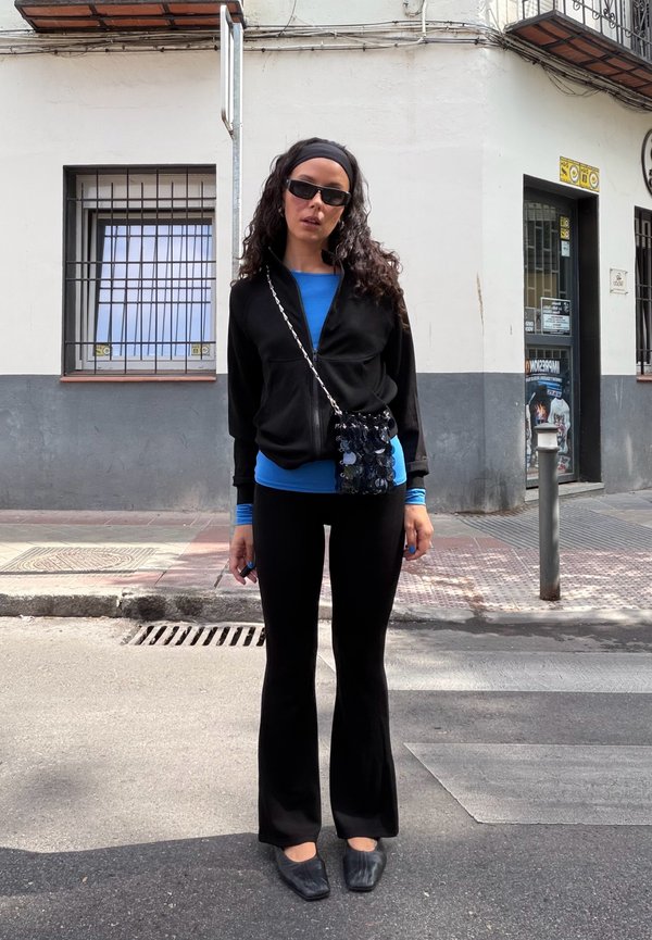 Mujer con cabello rizado, lleva gafas de sol negras, chaqueta negra, camiseta azul, pantalones negros y zapatos negros, está parada en una calle de la ciudad cerca de un edificio.