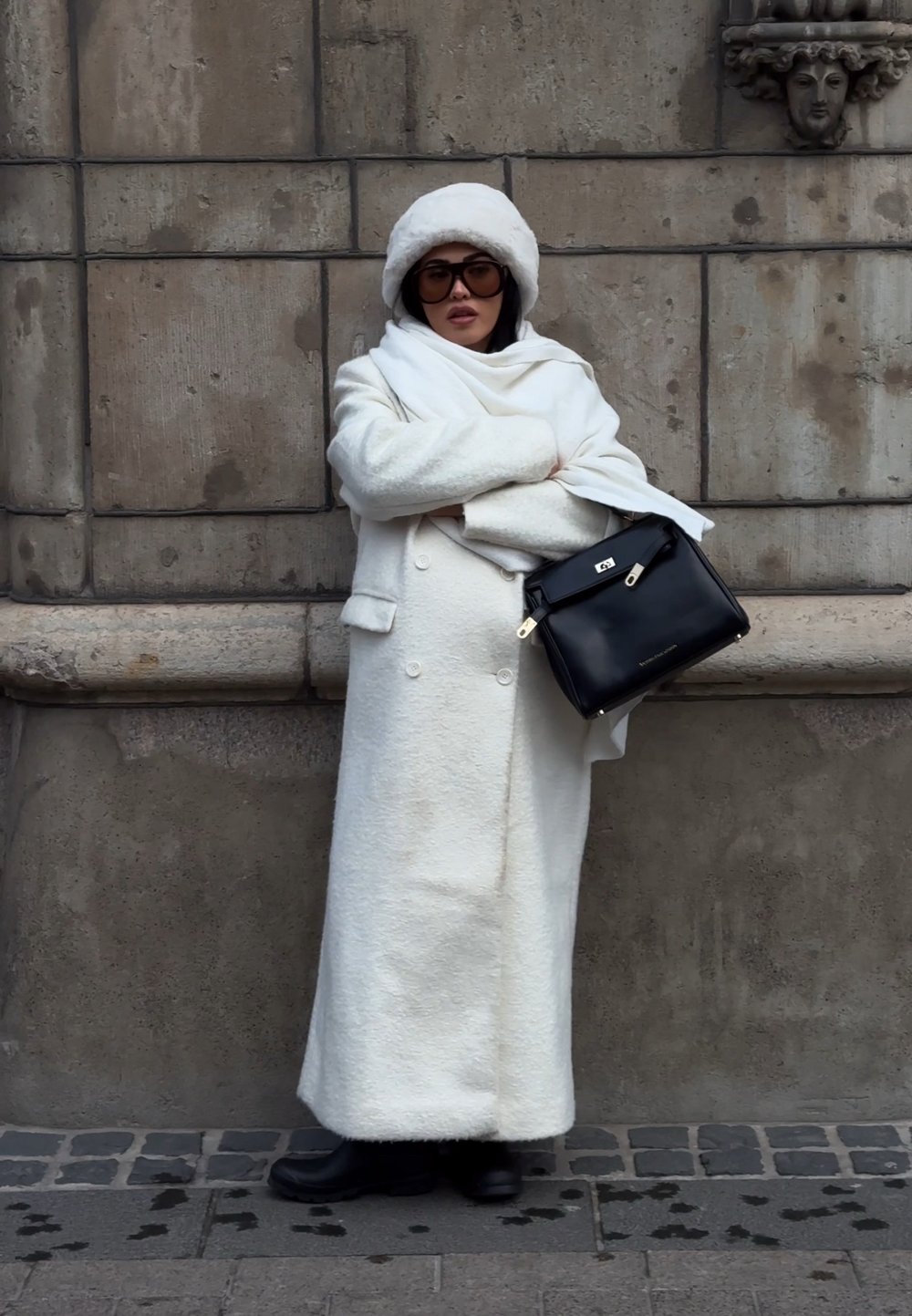 Femme portant un long manteau blanc, un chapeau blanc, des lunettes de soleil foncées et des bottes noires, se tient les bras croisés, tenant un sac à main noir près d'un mur en pierre.