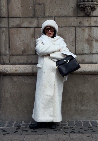 Mujer que lleva un abrigo largo blanco, un sombrero blanco, gafas de sol oscuras y botas negras, está de pie con los brazos cruzados, sosteniendo un bolso negro junto a una pared de piedra.