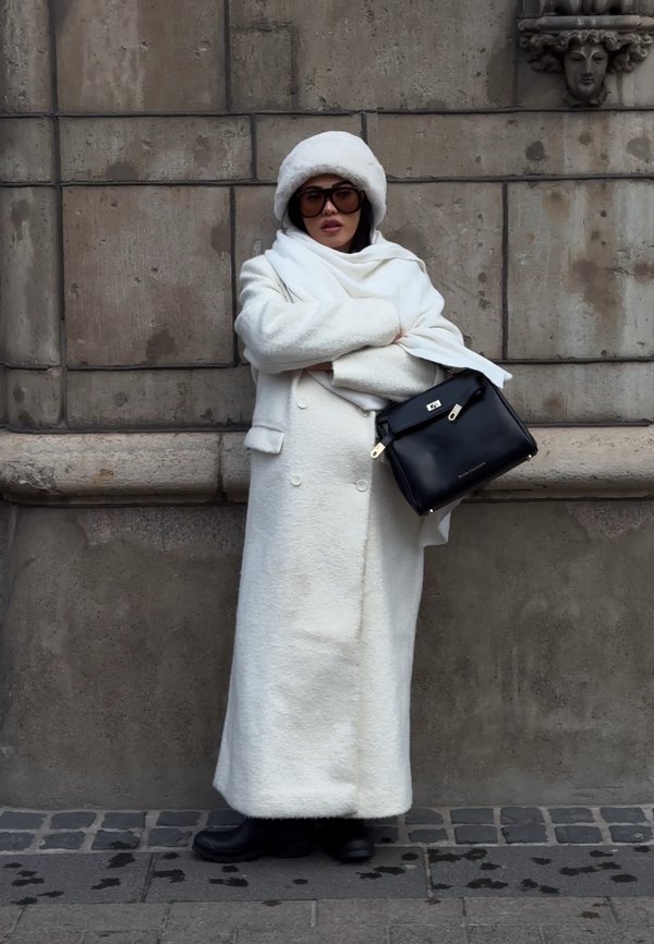 Žena v dlhom bielom kabáte, bielej čiapke, tmavých slnečných okuliaroch a čiernych čižmách stojí so založenými rukami a držiac čiernu kabelku pri kamennej stene.