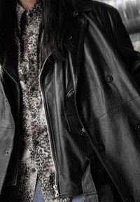 Veste en cuir noire à double boutonnage avec des boutons en forme d'étoile, superposée sur une chemise à boutons imprimée léopard. Détails de tissu texturé visibles.