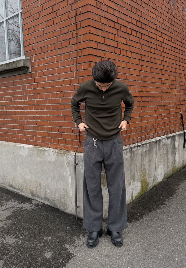 Osoba v tmavozelenej košeli s dlhým rukávom a voľných sivých nohaviciach s pripevnenými kľúčmi, stojaca na chodníku pri rohu červenej tehlovej steny.