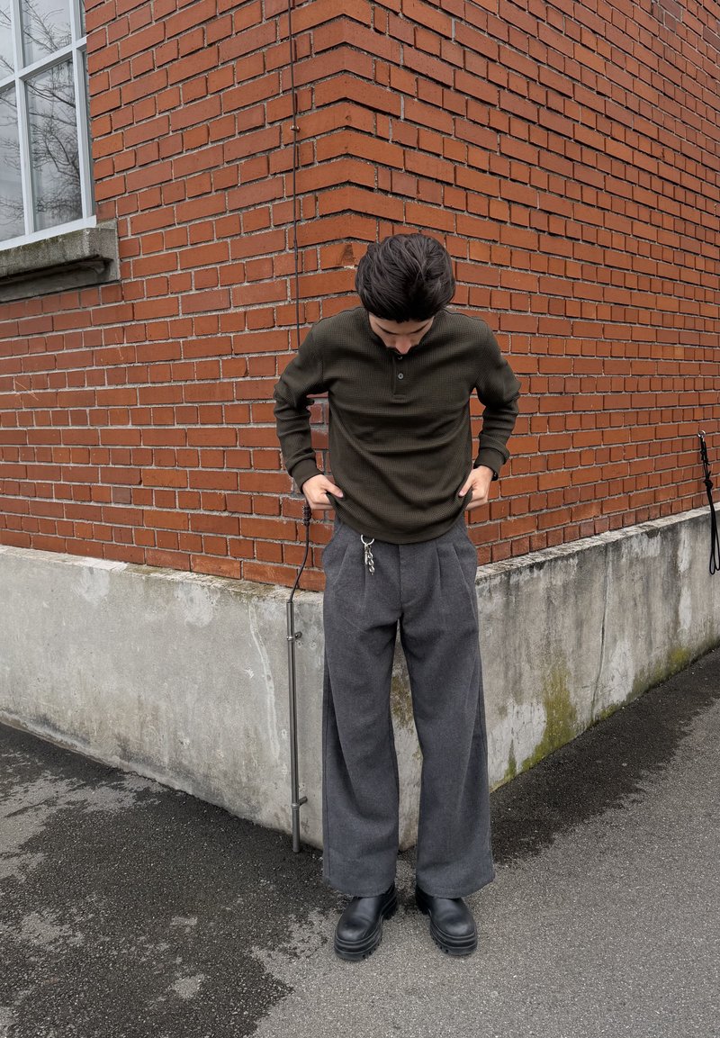 Osoba w ciemnozielonej koszuli z długim rękawem i luźnych szarych spodniach z zamocowanymi kluczami, stojąca na chodniku obok rogu czerwonej ceglanej ściany.