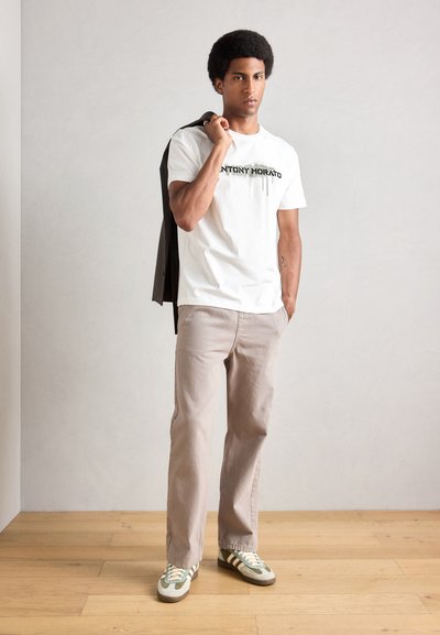 Camiseta blanca de algodón con texto impreso en negro, combinada con pantalones beige claro y zapatillas a rayas verdes. El modelo tiene un peinado afro.