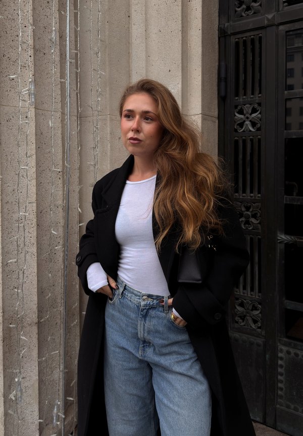 Svart oversized kåpe over en hvit langermet skjorte, lyseblå høytlivs jeans, med en teksturert steinvegg og en mørk metallør i bakgrunnen.