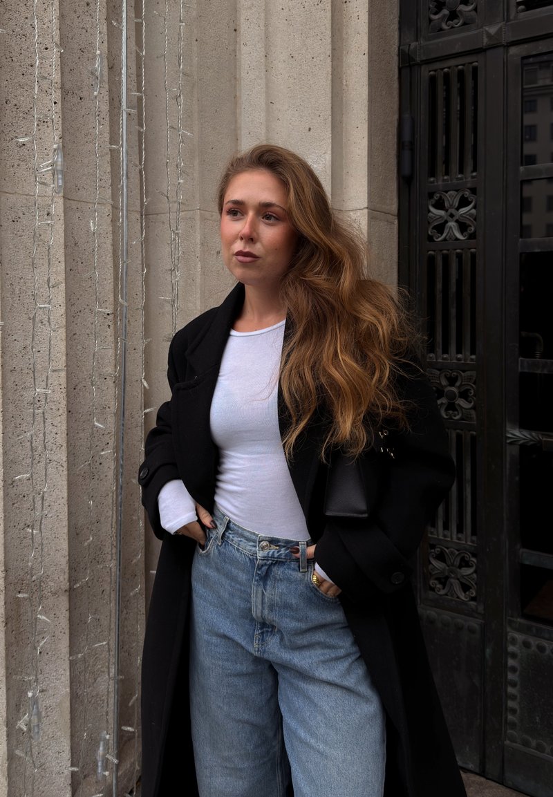 Musta ylisuurikokoinen takki valkoisen pitkähihainen paidan päällä, vaaleansiniset korkeavyötäröiset farkut, taustalla kiveksinen teksturoitu seinä ja tummasta metallista ovi.