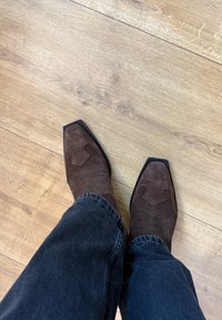 Hnedé semišové kovbojské topánky so špicatými špičkami, dekoratívnym prešitím na vrchu, nosené s tmavými riflemi na drevenej podlahe.