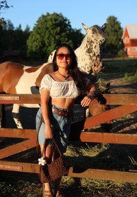 A person in a white crop top and denim shorts, holding a brown studded handbag, stands by a wooden fence with speckled horses in the background.