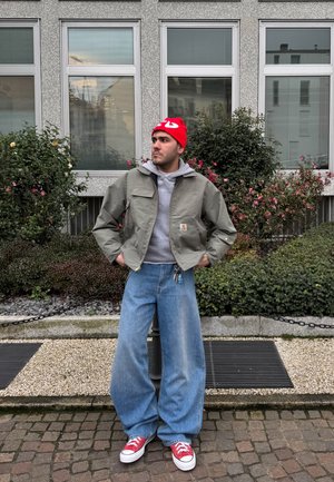 Zöld könnyű dzseki egy szürke kapucnis felső felett, széles kék farmernadrággal és piros tornacipővel párosítva. A piros sapka teszi teljessé a megjelenést.