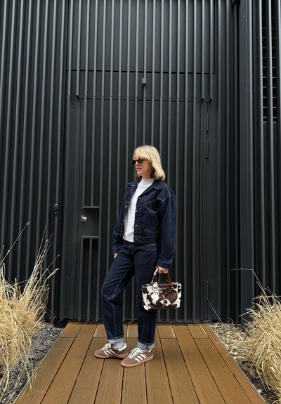 Chaqueta de mezclilla oscura y jeans con una camisa blanca, combinados con zapatillas marrones. Sosteniendo un pequeño bolso de estampado de vaca. Suelo de madera y piedra.