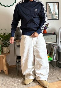 Personne portant une veste de survêtement Adidas bleu marine, un pantalon blanc à jambes larges et des chaussures beige, se tenant à l'intérieur près de plantes et d'une étagère avec des livres et une enceinte.