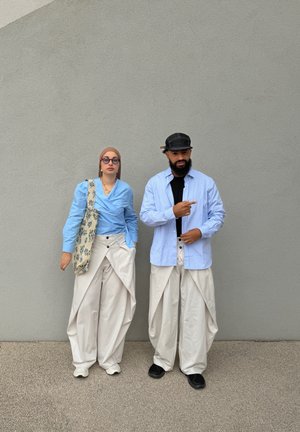 Dos personas están de pie contra una pared gris, vistiendo tops azul claro y pantalones blancos holgados; una lleva gafas y un pañuelo en la cabeza, la otra una gorra que apunta hacia la derecha.