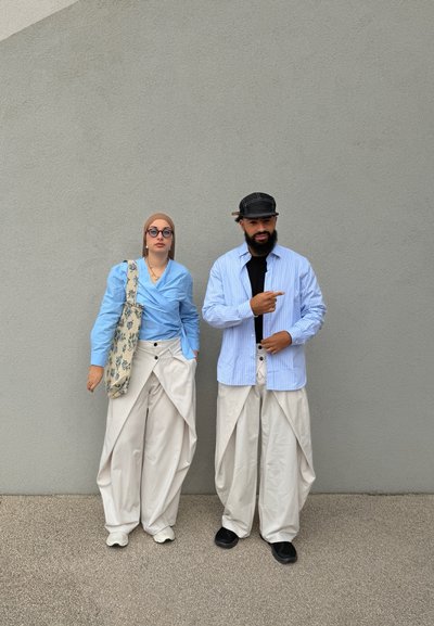 Dos personas están de pie contra una pared gris, vistiendo tops azul claro y pantalones blancos holgados; una lleva gafas y un pañuelo en la cabeza, la otra una gorra que apunta hacia la derecha.