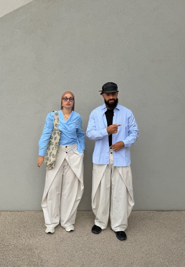 Dvaja ľudia stoja pri sivej stene, oblečení v svetlomodrých topoch a bielych voľných nohaviciach, jeden má okuliare a šatku na hlave, druhý čiapku a smeruje pravým smerom.