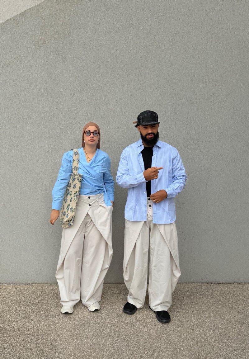 Kaksi henkilöä seisoo harmaan seinän edessä, yllään vaaleansiniset paidat ja valkoiset ylipitkät housut; toinen heistä on silmälasit ja huivi päässään, toinen lippalakki päässä ja osoittaa oikealle.