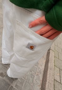 Biele bavlnené nohavice s veľkým bočným vreckom, s štvorcovou šedou etiketou a oranžovým logom. Čiastočne je viditeľný aj zelený rukáv.