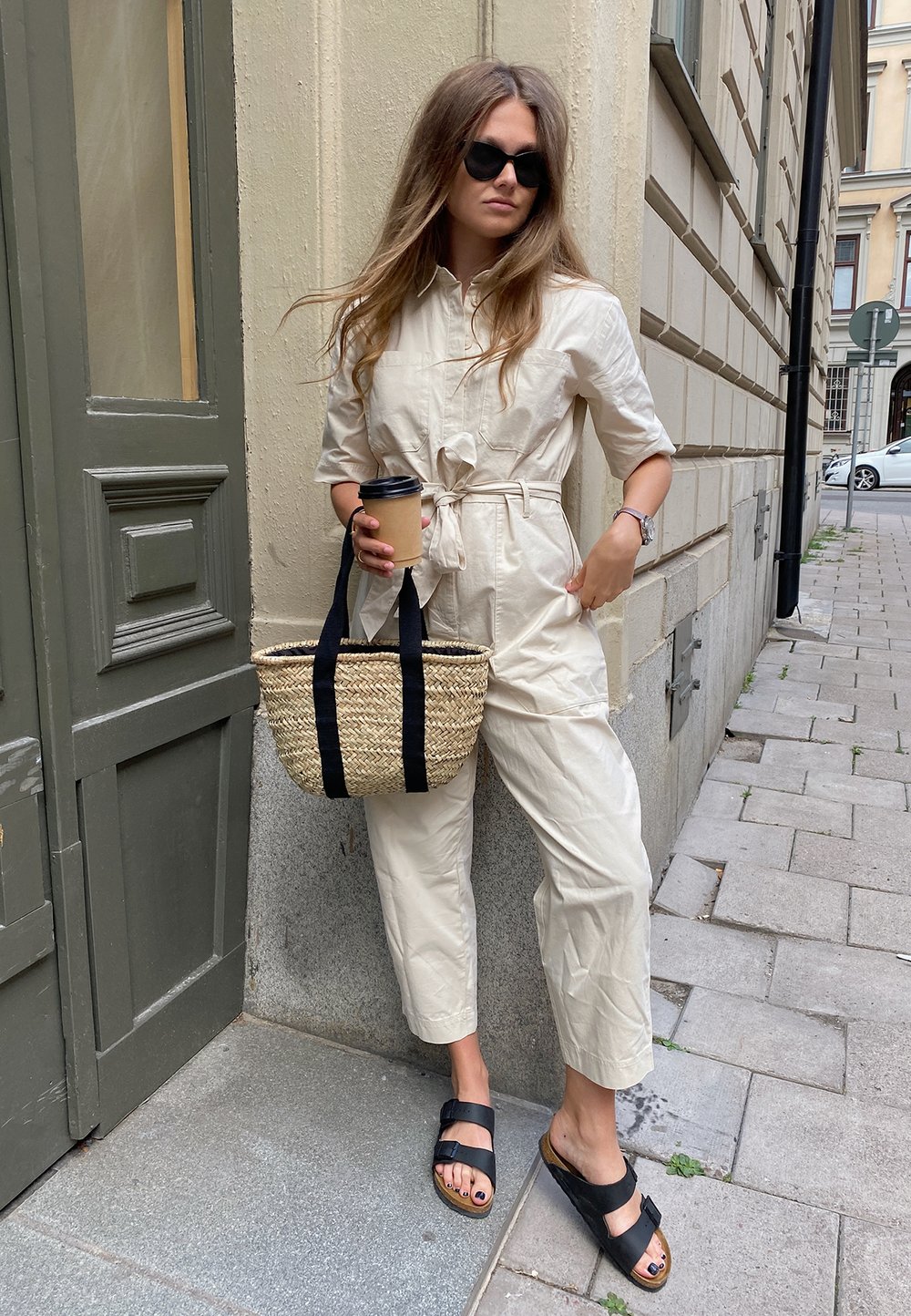 Világos bézs overál megkötős der�ékkal; fekete napszemüveg; szőtt szalma táska fekete pántokkal; fekete szandál; egy kőfal előtt áll.