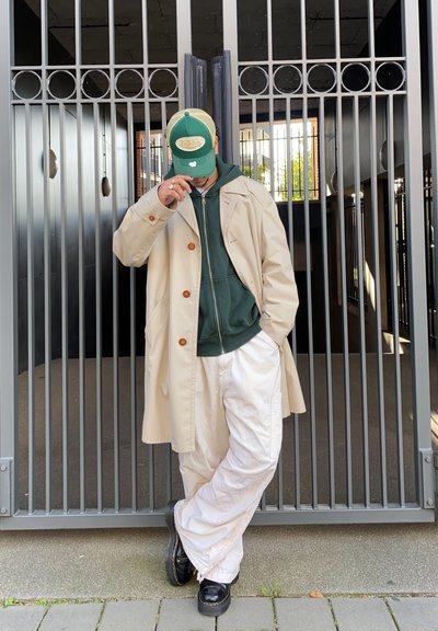 Abrigo trench beige con botones marrones, sudadera verde con capucha, pantalones blancos de corte holgado y calzado negro. El fondo presenta una puerta de metal.