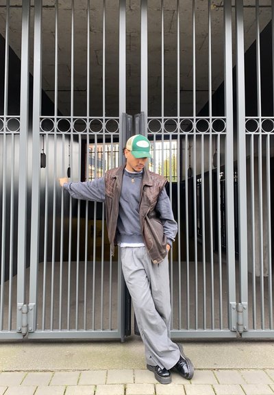 Chaleco de cuero marrón sobre sudadera gris, pantalones de pierna ancha grises y zapatos negros. De pie contra una puerta de metal gris con barras verticales.