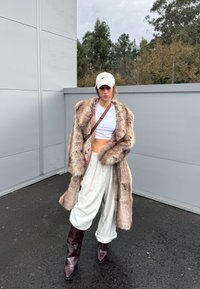 Femme portant un crop top blanc, un manteau surdimensionné en fausse fourrure, un pantalon ample gris clair, des bottes marron foncé et une casquette Nike blanche, debout en plein air.