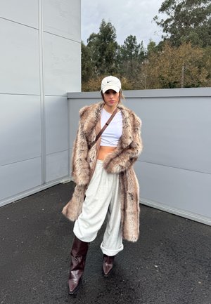 Mujer con top corto blanco, abrigo de piel sintética oversize, pantalones sueltos gris claro, botas marrón oscuro y gorra blanca de Nike, de pie al aire libre.