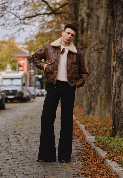 Chaqueta bomber de cuero marrón con un cuello de borreguillo, suéter crema, corbata estampada y pantalones acampanados negros. Entorno urbano con hojas de otoño.