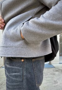 Egy szürke dzsekit viselő személy, aki a zsebeibe dugott kezekkel sötét farmernadrágban áll egy épület bejárata közelében.