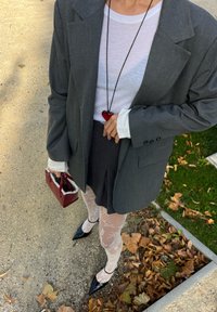 Grå blazer over en hvid ribbet skjorte, sort nederdel, mønstrede hvide strømpebukser, sorte spidse sko og en rød håndtaske. Hjertevedhæng detalje.