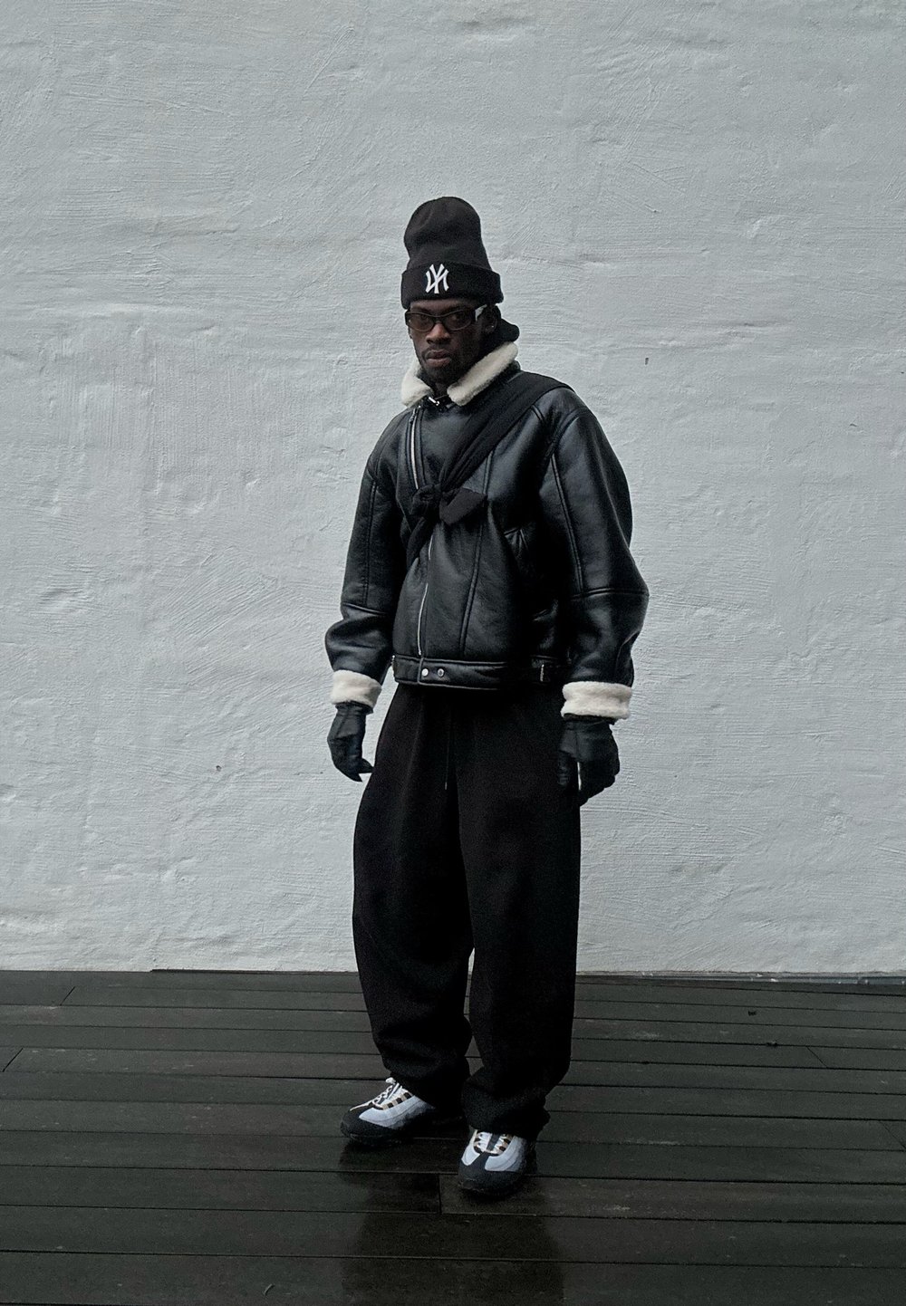 Giubbotto bomber in pelle nera con colletto in pelliccia bianca, guanti neri, pantaloni neri oversize e sneakers grigie, su uno sfondo grigio chiaro.