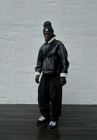 Giubbotto bomber in pelle nera con colletto in pelliccia bianca, guanti neri, pantaloni neri oversize e sneakers grigie, su uno sfondo grigio chiaro.