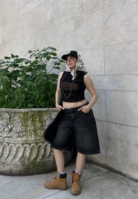 Črn brezrokavnik, oversize črni denim kratke hlače, rjavi delovni škornji in kapa s potiskom v slogu bandane, ki stoji ob okrasnem cvetličnem loncu.