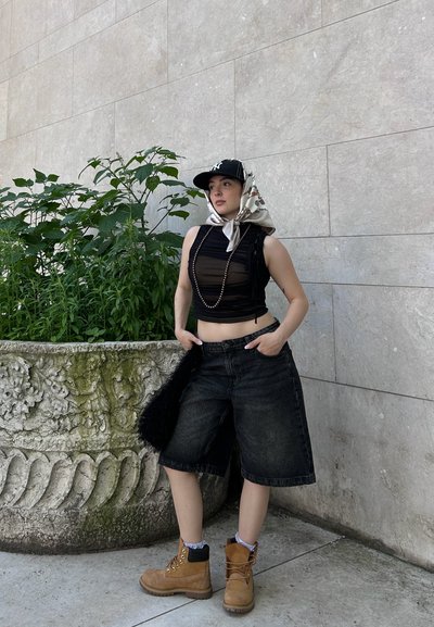 Top sin mangas negra, pantalones cortos de mezclilla negros de gran tamaño, botas de trabajo beige y una gorra tipo pañuelo estampada, de pie cerca de un macetero decorativo.