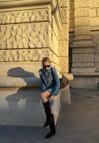 Traper jakna i haljina, smeđe čizme do koljena, uzorčana torba, stoji pored kamenog zida s teksturom, blago poslijepodnevno svjetlo, duga sjena.