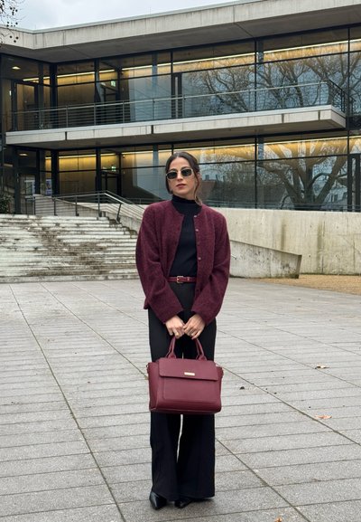 Mujer con gafas de sol, un cárdigan burdeos, un conjunto negro, sosteniendo un bolso burdeos, de pie en una zona pavimentada frente a un edificio moderno.