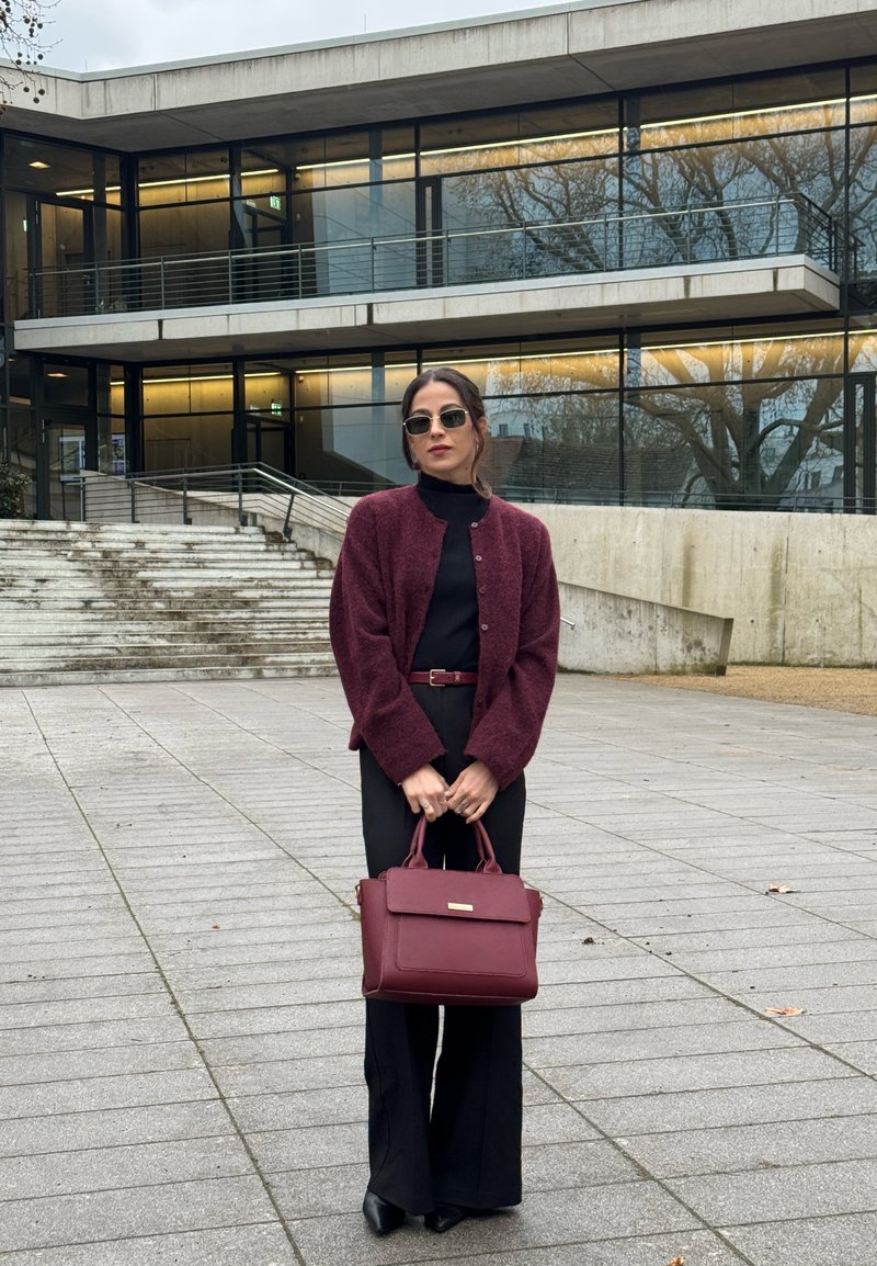 Γυναίκα με γυαλιά ηλίου, μπορντό ζακέτα, μαύρο σύνολο, κρατώντας μια μπορντό τσάντα, στέκεται σε πλακόστρωτη περιοχή μπροστά από ένα μοντέρνο κτίριο.