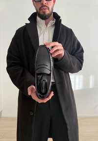 Άνδρας με μαύρο παλτό και λευκό πουκάμισο κρατά μαύρο δερμάτινο παπούτσι τύπου loafer με ετικέτα Tommy Hilfiger προς την κάμερα.