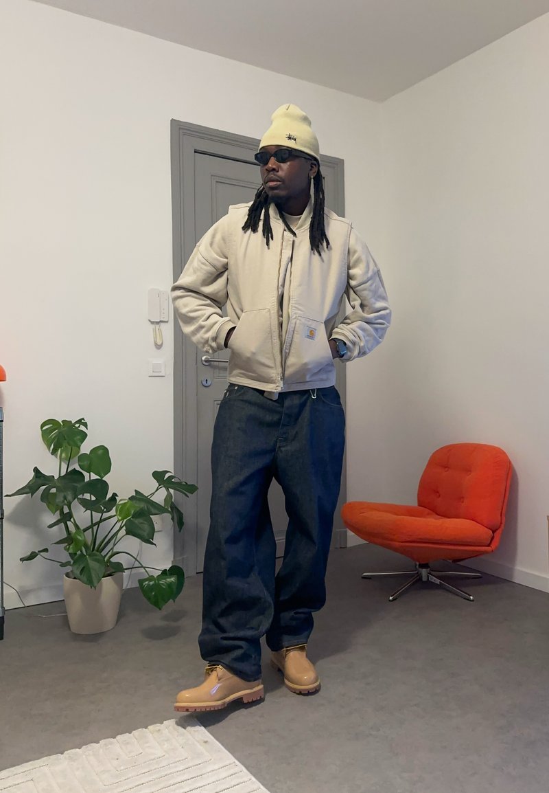 Beige bomberjacka, lösa mörka denimbyxor, bruna stövlar, gul mössa, solglasögon. Inomhusmiljö med en grön växt och en orange stol.