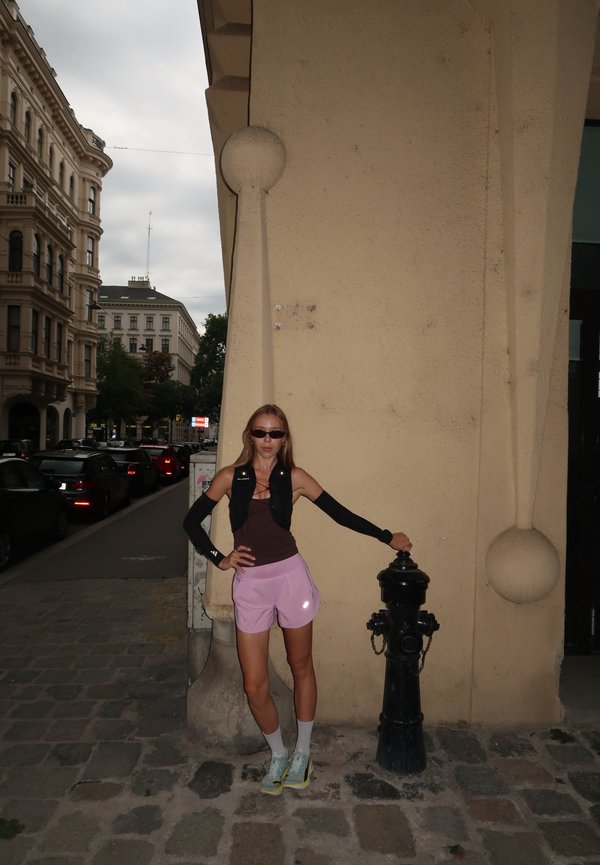 Femme en tenue sportive appuyée sur une bouche d'incendie noire dans une rue pavée en zone urbaine avec des bâtiments historiques et des voitures garées.