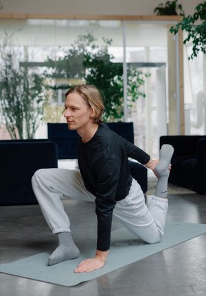 Un homme en chemise noire à manches longues et en pantalon de survêtement gris effectue un étirement de yoga sur un tapis gris clair dans un espace intérieur moderne.