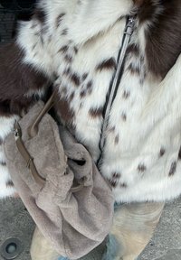 Asmuo, vilkintis baltai rudos dėmėtos kailinės striukės su sidabriniu užtrauktuku, laiko minkštą taupe spalvos tekstūruotą maišą su odiniais diržais.