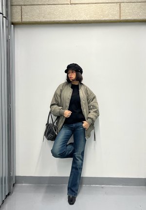 Persona con chaqueta bomber verde, camiseta negra, vaqueros azules, sombrero negro y botas, de pie contra una pared blanca lisa con una pierna doblada.